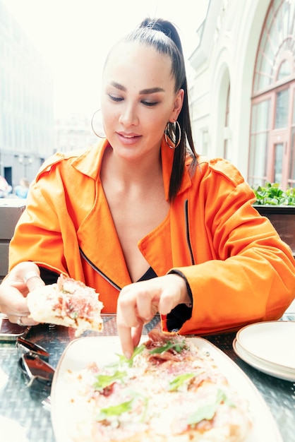 Uma jovem elegante senta-se em um café da cidade e come pizza Sorrindo linda morena em uma jaqueta laranja brilhante Exposição vertical ao sol