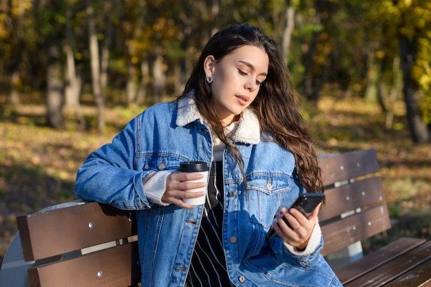 Uma jovem em um banco no parque outono com café e um smartphone nas mãos