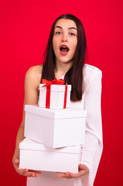 Foto uma jovem em um vestido de noite segura muitas caixas com presentes para o natal um modelo surpreso em um fundo vermelho posa no estúdio e olha para a câmera