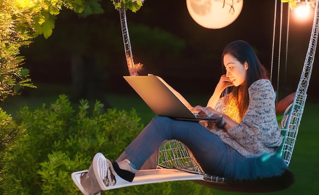 Uma jovem em uma cadeira de balanço de jardim com seu laptop iluminado pela luz suave da lua