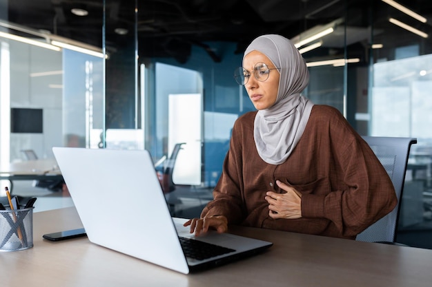 Uma jovem empresária árabe em um hijab está sentada em uma mesa no escritório e sofrendo de estômago