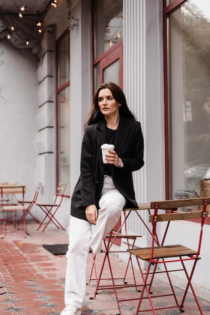 Uma jovem empresária confiante com uma xícara de café está posando do lado de fora do café no terraço Uma garota alegre usa um estilo de negócios casual que está posando perto do café