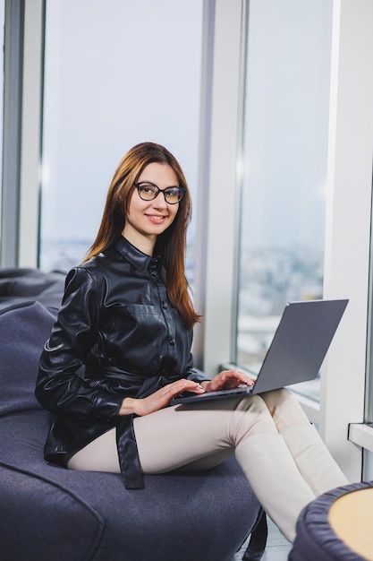 Uma jovem empresária está sentada enquanto trabalha em um laptop O conceito de uma mulher moderna e bem-sucedida Jovem garota curvilínea séria de óculos sentada em uma cadeira em um espaço aberto de trabalho moderno