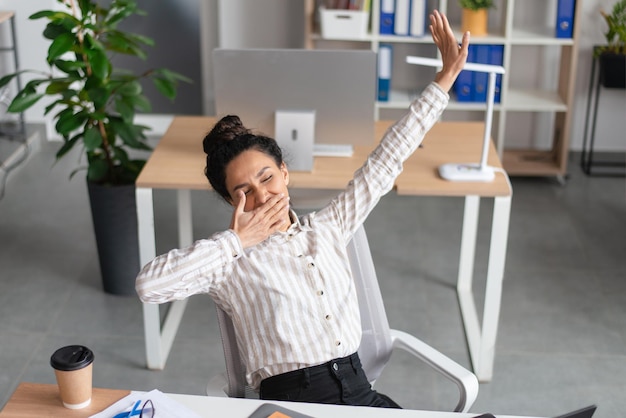 Uma jovem empresária sonolenta bocejando e se esticando cansada de trabalhar no escritório sentindo-se sonolenta em