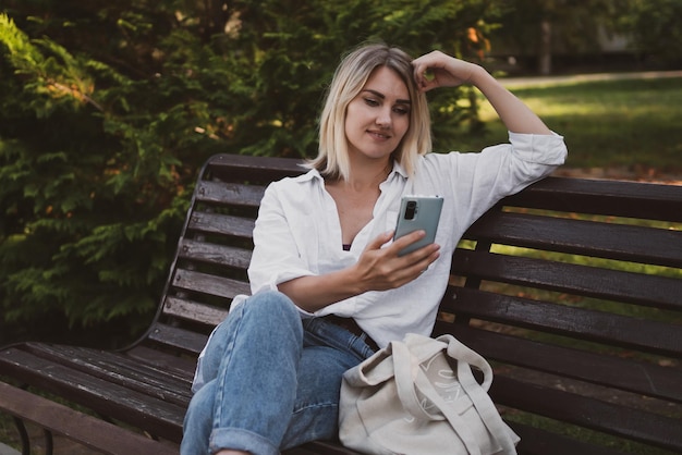 Uma jovem encantadora em roupas casuais senta-se em um banco no parque e usa um telefone celular para se comunicar