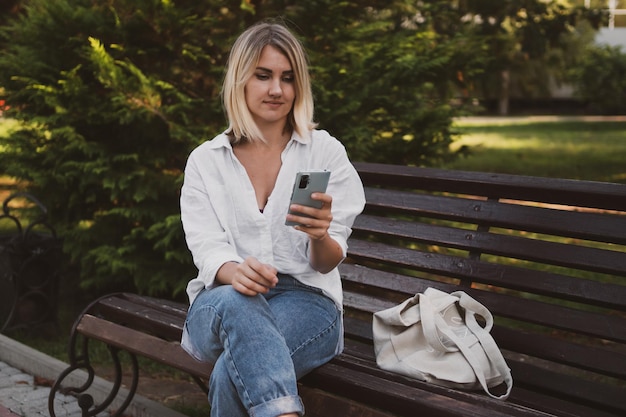 Uma jovem encantadora em roupas casuais senta-se em um banco no parque e usa um telefone celular para se comunicar