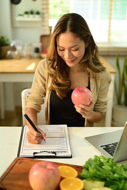 Uma jovem encantadora segurando uma maçã e escrevendo uma receita na mesa com frutas frescas Dieta nutrição correta alimentação saudável