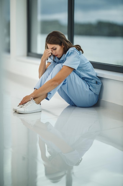 Uma jovem enfermeira estressada sentada no chão perto de uma janela em um corredor de hospital vazio durante a pandemia de Covid-19.