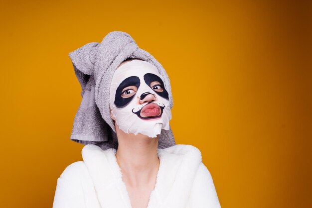 Uma jovem engraçada com uma toalha na cabeça e uma túnica branca cuida de si mesma no rosto uma máscara com cara de panda