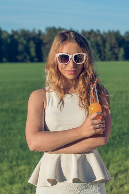 Uma jovem esbelta de óculos com cabelos cacheados sorri e bebe um coquetel alcoólico ou não alcoólico através de um canudo de uma garrafa em um dia ensolarado de verão e sorri