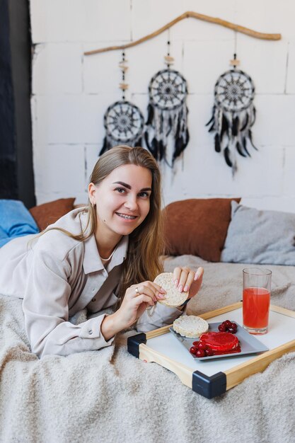 Uma jovem esbelta está sentada na cama e tomando um delicioso café da manhã com bolos de arroz e suco fresco Alimentação Saudável