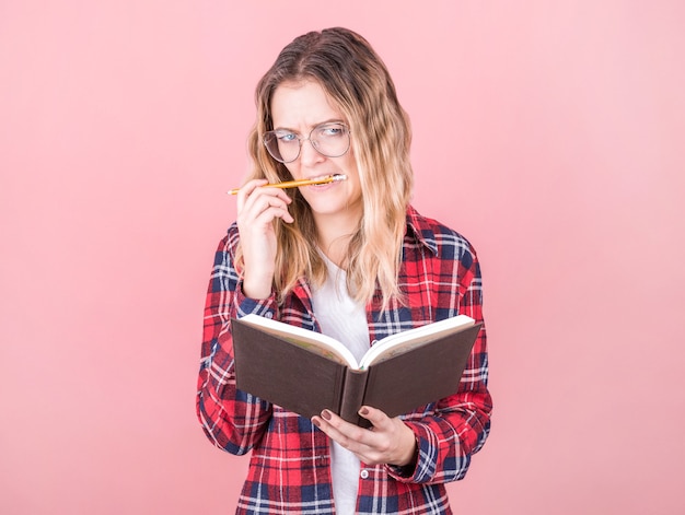 Uma jovem escritora de cabelos loiros com uma camisa xadrez segura um livro nas mãos com um mal-entendido, roe um lápis e pensa no que escrever. dia internacional dos escritores