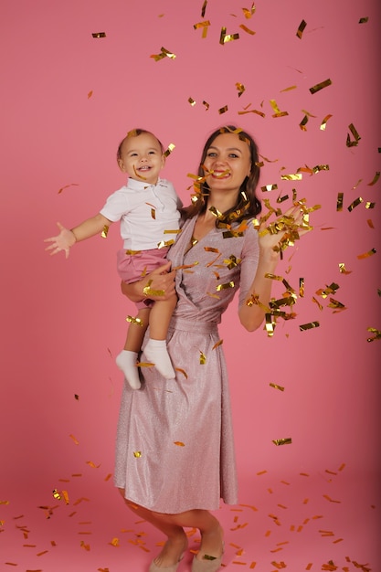 uma jovem esguia em um vestido rosa segura seu filho nas mãos pegando confete