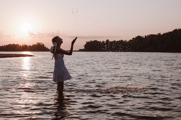 Uma jovem espirra água. foto de uma loira esguia, linda e feliz em um vestido de verão e um chapéu de palha em pé no rio contra o pôr do sol e levantando as mãos.