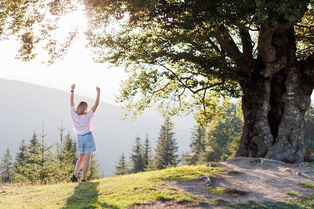 Uma jovem está de pé perto de uma grande árvore em uma colina e levantou as mãos nas montanhas dos Cárpatos