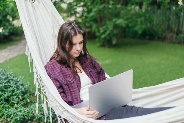 Uma jovem está deitada em uma rede com um laptop no jardim e trabalha remotamente