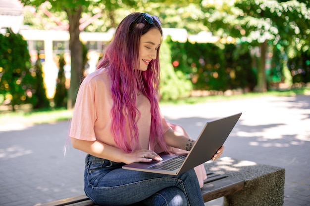 Uma jovem está estudando no parque da primavera, sentada no banco de madeira e navegando em seu laptop