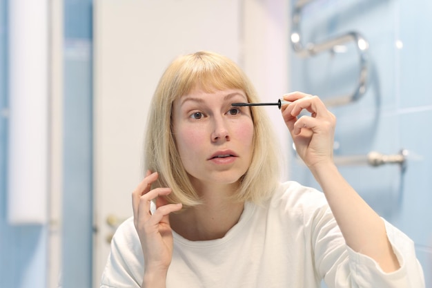 Uma jovem está fazendo maquiagem. Menina pinta os cílios com rímel, olhando seu reflexo no espelho na parede. Conceito de procedimentos de maquiagem feminina de manhã.