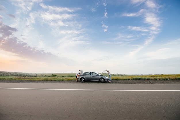 Uma jovem está perto de um carro quebrado no meio da rodovia durante o pôr do sol e tenta consertá-lo. Avaria e reparação do carro. Resolvendo o problema.