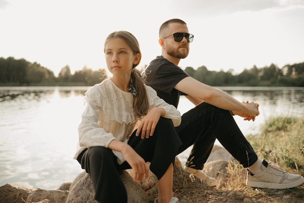 Uma jovem está posando em pedras com o pai com barba e óculos de sol na costa do lago. família elegante de pai solteiro de férias no pôr do sol.