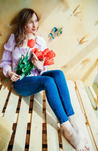Uma jovem está segurando um buquê de tulipas vermelhas e sentado em um pódio de madeira o conceito de 8 de março dia dos namorados um presente maravilhoso para uma menina retrato de primavera de uma mulher