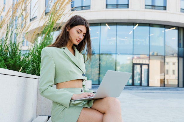 Uma jovem está sentada em um banco com um laptop e trabalhando do lado de fora empresária elegante na rua