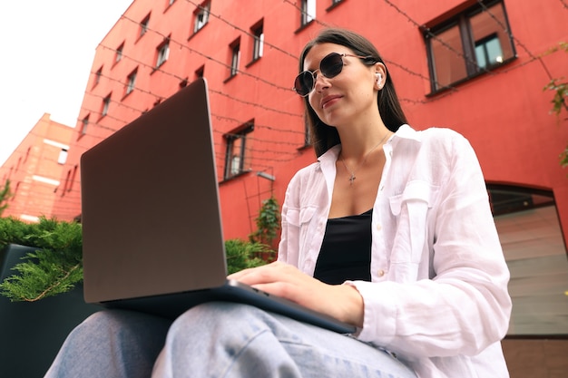 Uma jovem está sentada em um banco e trabalhando com um laptop na rua