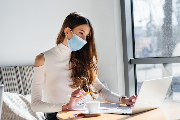 Uma jovem está sentada em um café usando uma máscara e trabalhando em um computador