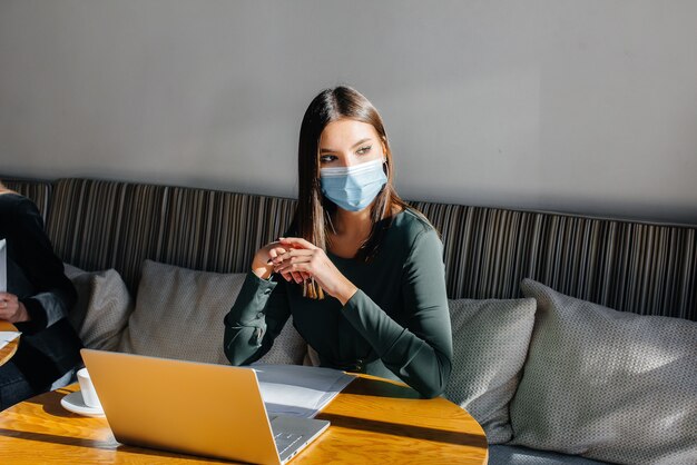 Uma jovem está sentada em um café usando uma máscara e trabalhando em um computador
