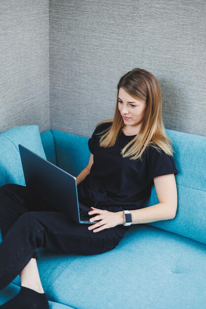 Uma jovem está sentada em um sofá e trabalhando em um laptop Trabalho remoto Trabalho em casa online