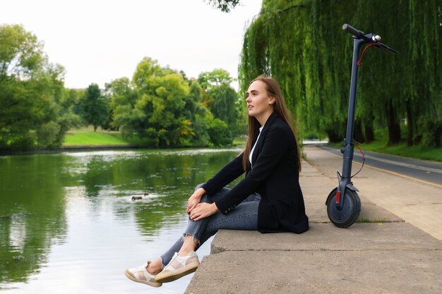 Uma jovem está sentada na margem do rio ao lado de uma scooter elétrica