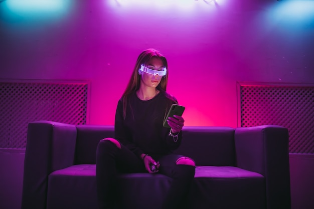 Uma jovem está sentada no sofá usando óculos de néon e usando um gadget de foto de alta qualidade