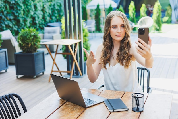 Uma jovem está trabalhando em um laptop e falando ao telefone em um aconchegante local de trabalho ao ar livre no terraço de um café O conceito de trabalho remoto em um café confortável ou trabalho durante as férias