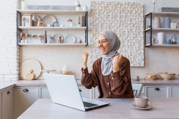 Uma jovem estudante latino-americana feliz em hijabsits em casa na frente de um laptop levantando-a