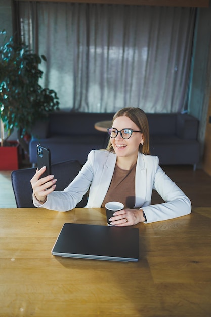 Uma jovem feliz com longos cabelos loiros em roupas elegantes está sentada em uma mesa com um laptop desfrutando de uma xícara de café fresco e falando ao telefone Trabalho em um escritório moderno com grandes janelas