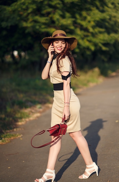Uma jovem feliz com um chapéu de aba larga caminha no parque no verão com uma bolsa e fala ao telefone