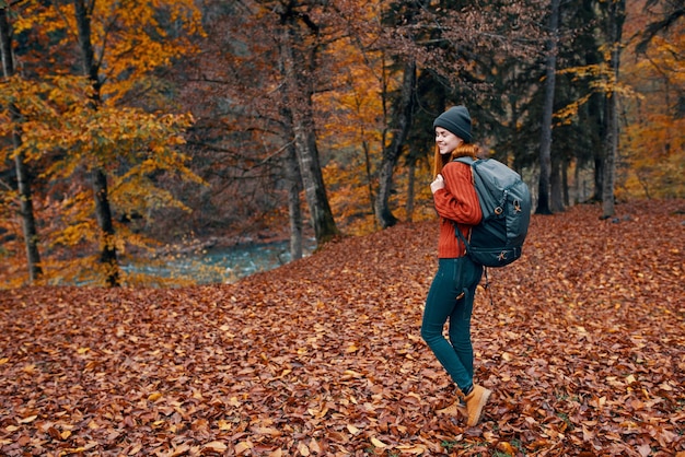 Uma jovem feliz com uma mochila em botas jeans e um suéter estão caminhando na floresta de outono perto das árvores altas