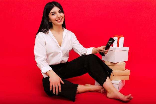 Uma jovem feliz de camisa e jeans está sentada ao lado de presentes de Natal em um fundo vermelho O modelo segura uma taça de champanhe