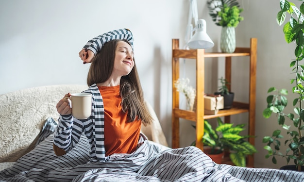 Uma jovem feliz de pijama está sentada na cama com uma xícara de café aromático e se espreguiçando depois de acordar