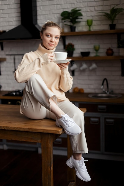 Uma jovem feliz está em um ambiente aconchegante em casa, senta-se à mesa da cozinha e segura uma xícara de café
