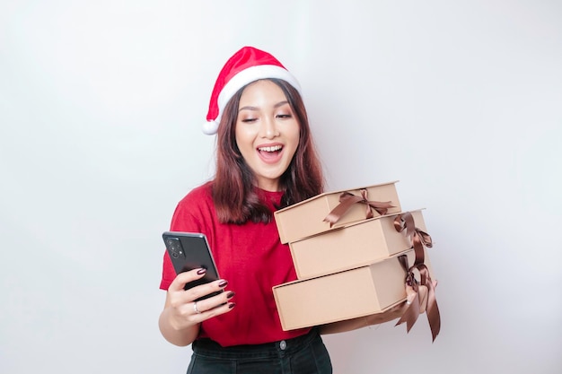 Uma jovem feliz está usando chapéu de Papai Noel39 enquanto segura presentes de Natal e seu smartphone