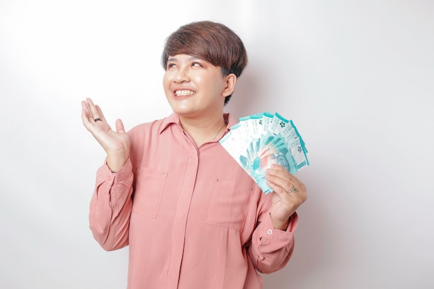 Uma jovem feliz está vestindo camisa rosa e segurando dinheiro em rupia indonésia isolada por fundo branco