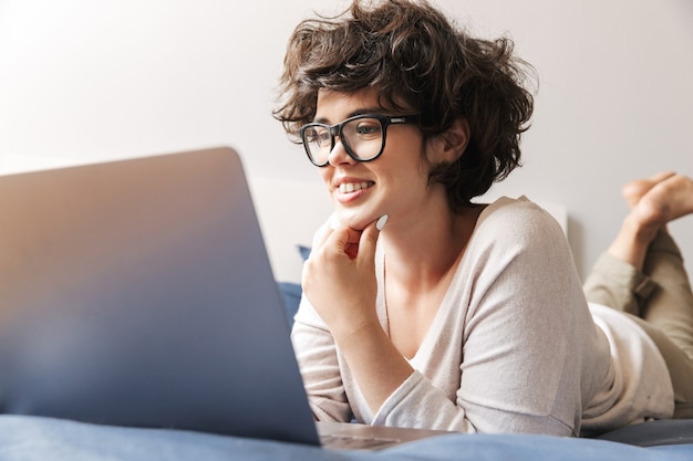 Uma jovem feliz fica dentro da cama, usando o computador portátil.