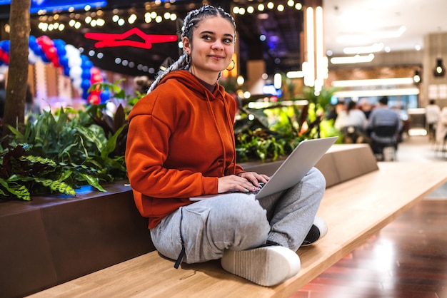 Uma jovem feliz sentada no banco com as pernas cruzadas usando seu laptop em um shopping center