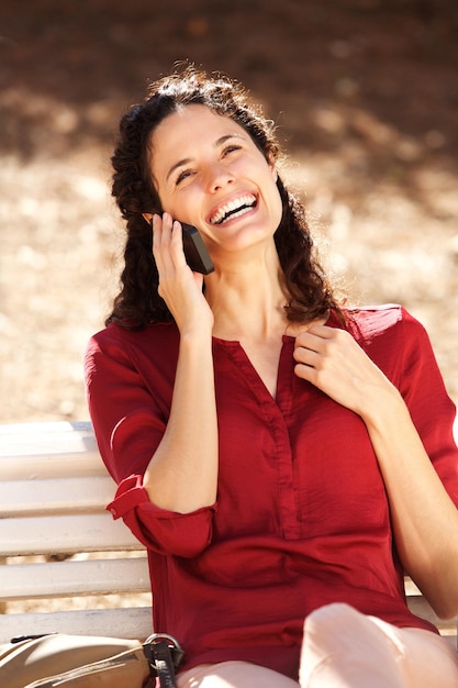 Uma jovem feliz sentada num banco do parque a falar ao telemóvel.