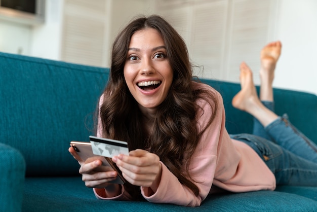 Uma jovem feliz surpresa dentro de casa em casa usando telefone celular, segurando o cartão de crédito no sofá.