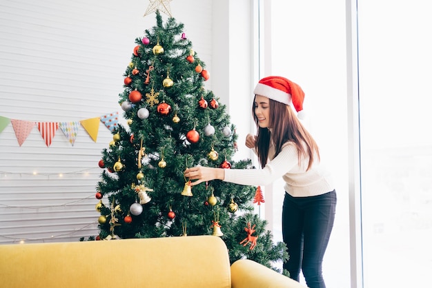 Uma jovem feliz usando um chapéu e suéter de Papai Noel sorri e gosta de decorar várias bugigangas na árvore de Natal na sala de estar em casa Desfrutando e celebrando o feriado de Natal