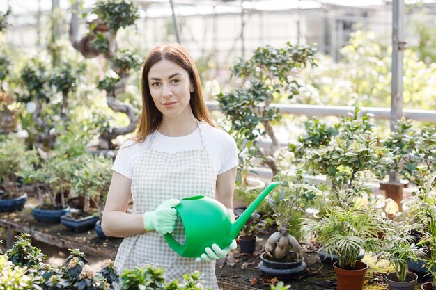 Uma jovem fica no meio de uma grande estufa segurando um regador e sorrindo para a câmera O conceito de cuidar de plantas de casa