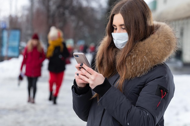 Uma jovem fica perto da estrada e escreve uma mensagem no smartphone em uma máscara médica Proteção contra surtos de vírus gripe e poluição do ar de carros na cidade