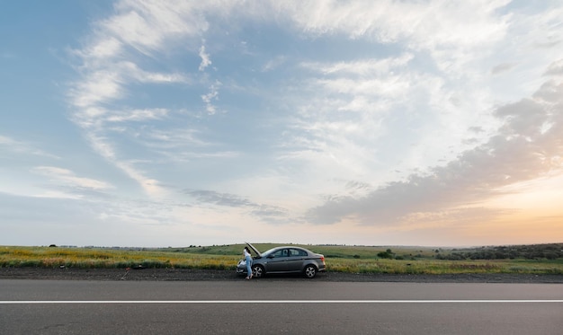 Uma jovem fica perto de um carro quebrado no meio da estrada durante o pôr do sol e tenta pedir ajuda no telefone Esperando por ajuda Serviço de carro Avaria do carro na estrada